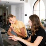 Two women engaged in a collaborative discussion at a modern office setting over laptops.