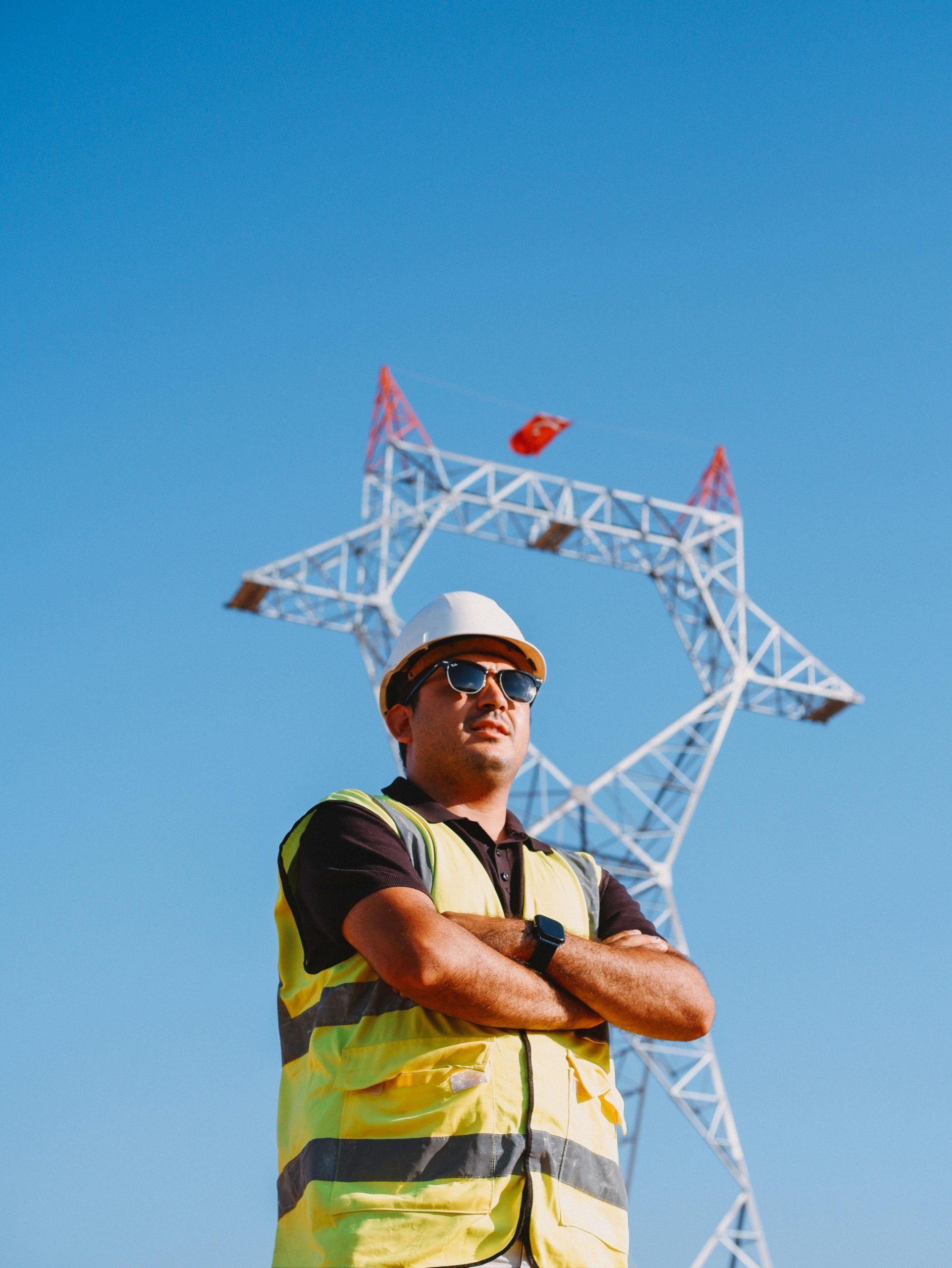 Confident construction worker in safety gear stands before a power line tower against a blue sky.